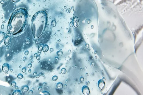 Gros plan d'un gel translucide, de couleur bleue, avec de nombreuses bulles d'air à l'intérieur, étalé sur une surface lisse et blanche. Le gel a une texture brillante et luisante et semble légèrement visqueux.