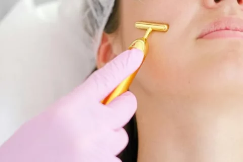 Une personne portant un filet à cheveux reçoit une injection cosmétique pour le visage tandis qu'une autre main gantée utilise un outil de massage en or sur sa joue.