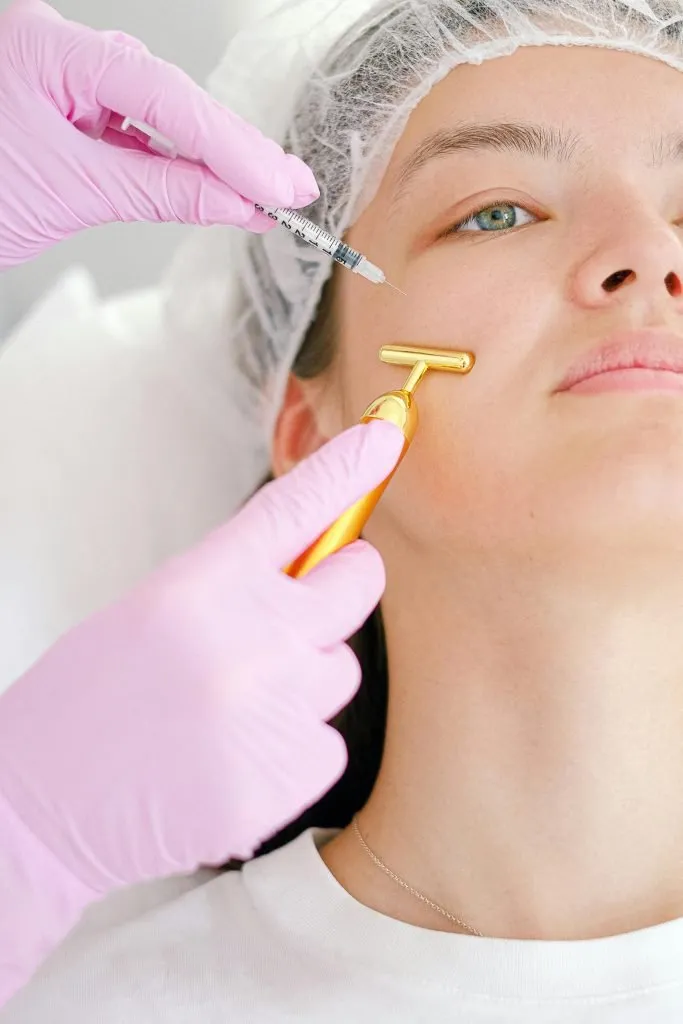 Une personne portant une charlotte reçoit une intervention cosmétique sur le visage, une main gantée injectant une seringue près de la joue et une autre utilisant un outil de massage facial en or sur le bas du visage.