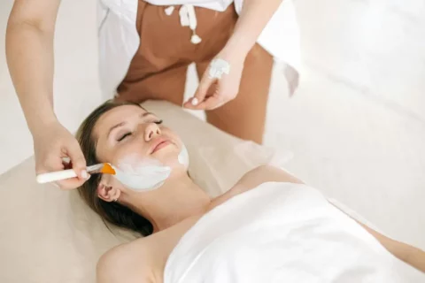 Une femme est allongée sur un lit de spa, les yeux fermés, tandis qu'une autre personne lui applique un masque facial blanc à l'aide d'une brosse. L'atmosphère est calme et relaxante.