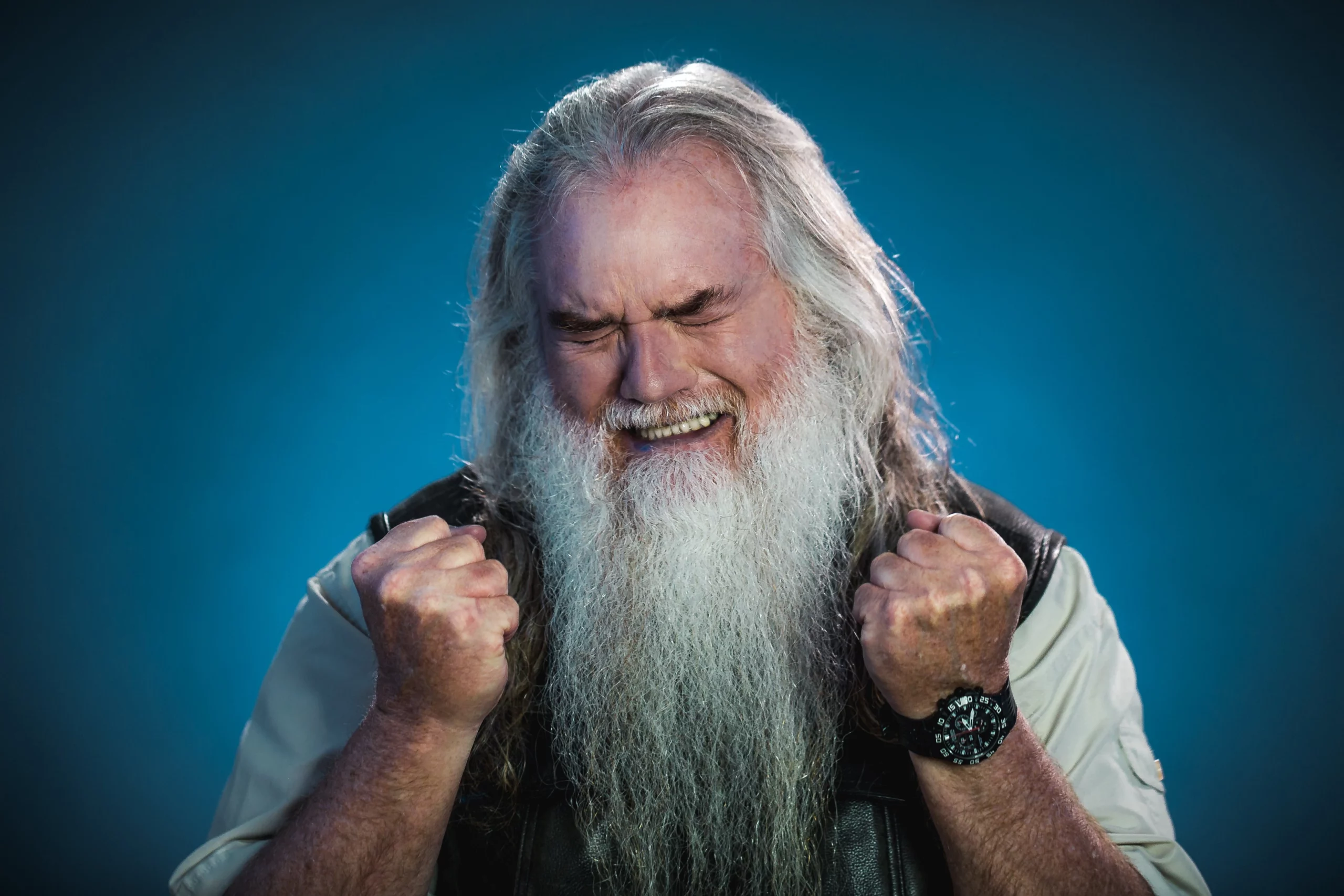 Un homme âgé aux longs cheveux gris et à l'épaisse barbe blanche serre les poings et sourit joyeusement, les yeux fermés, sur un fond bleu.